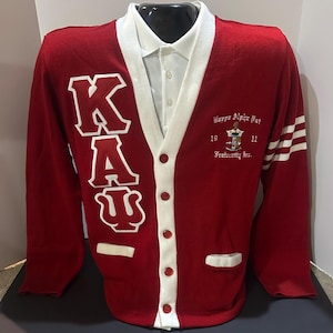 May include: Red cardigan sweater with white trim and buttons. The sweater has a large white "KAU" embroidered on the front and a Kappa Alpha Psi Fraternity Inc. logo on the right side.