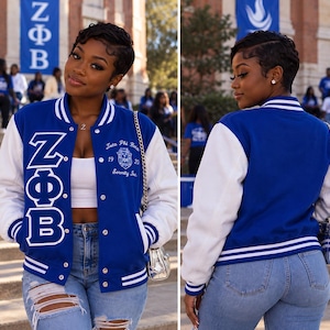 May include: A royal blue and white varsity jacket featuring the Greek letters ZΦB embroidered in white on the front left panel. The jacket includes a white embroidered crest with the text Zeta Phi Beta Sorority Inc. 1920 on the front right.