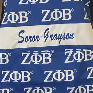 May include: A blue apron with a white stripe and the text "Soror Grayson" embroidered on it. The apron is covered in a repeating pattern of the letters "Z&Phi;&Beta;" in white.