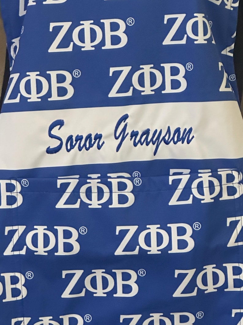 May include: A blue apron with a white stripe and the text "Soror Grayson" embroidered on it. The apron is covered in a repeating pattern of the letters "Z&Phi;&Beta;" in white.