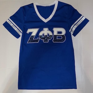 May include: A royal blue jersey-style t-shirt with a white V-neck and white stripes on the sleeves. The shirt features the letters "ZΦB" in white and navy blue glitter.
