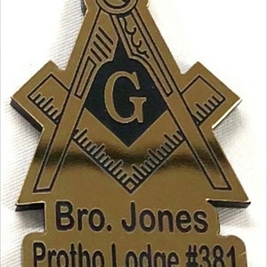 May include: Gold-coloured Masonic pin with a black square and compass design, a black letter G in the centre, and the text "Bro. Jones Protho Lodge #381".