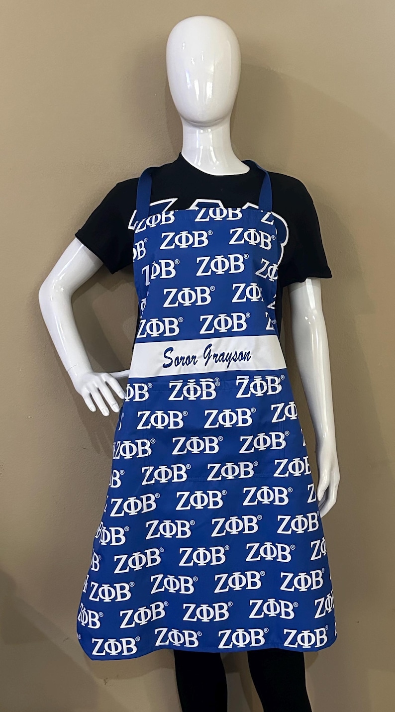 May include: A blue and white apron with the letters "Z&Phi;&Beta;" repeated all over it. The apron has a white pocket with the text "Soror Grayson" written on it.