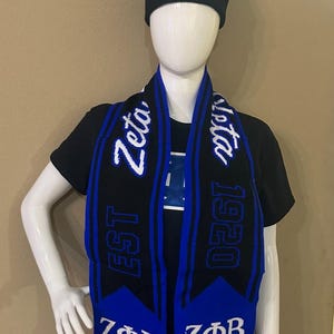 May include: Black knit beanie and scarf set with blue and white embroidered text. The beanie has a pom-pom and the scarf features the text "Zeta" and "1920". The set is displayed on a mannequin.