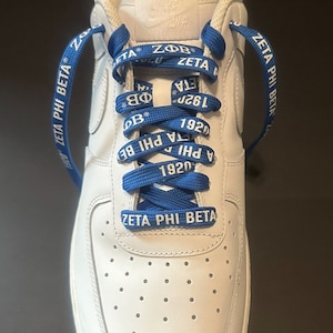 May include: White leather sneakers with blue shoelaces that have the text "Zeta Phi Beta" and "1920" printed on them.