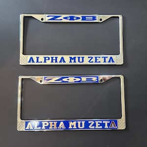 以下が含まれることがあります： 青と金の文字が書かれた銀色のナンバープレートフレームが2つ。上のフレームには「ZΦB」と「ALPHA MU ZETA」と書かれている。下のフレームには同じことが書かれている。