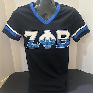Op de afbeelding: Zwart V-hals T-shirt met blauwe en witte accenten. Het shirt heeft de letters "ZΦB" in wit met blauwe glitter. De mouwen hebben blauwe en witte strepen. Het shirt wordt getoond op een zwarte mannequin.