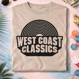 May include: A beige t-shirt with a black graphic that reads "WEST COAST CLASSICS" above a stylized record design. Sunglasses, play money, and cocktail umbrellas are scattered around the shirt.