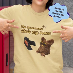 May include: A light yellow t-shirt with the text "The Government Stole My Beans" in brown letters with a retro font. A brown furry toy with yellow eyes is on the right side of the shirt. A black wallet with a pink interior is on the left side of the shirt. The text "Because Normal Shirts Are Boring" is in a speech bubble in the top right corner of the image.