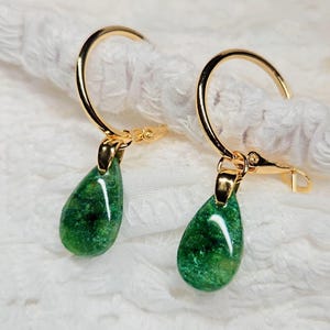 May include: A pair of gold-toned hoop earrings featuring dangling, teardrop-shaped pendants. The pendants are a deep, shimmering emerald green with a glossy, polished finish. The gold hoops have a simple, circular design with a secure clasp mechanism.