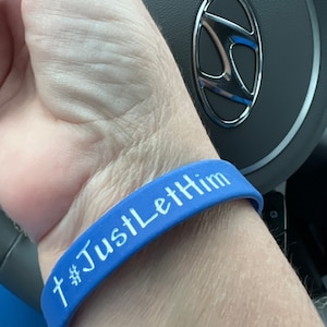 May include: A blue silicone wristband with the text "#JustLetHim" and a cross symbol in white. The wristband is worn on a wrist, with a car steering wheel in the background.
