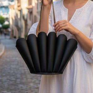 May include: A black handbag with a unique, fan-like design. The bag has a structured shape with a series of curved, vertical panels. It features thin, dark straps and gold-colored hardware. The bag is held against a blurred background.