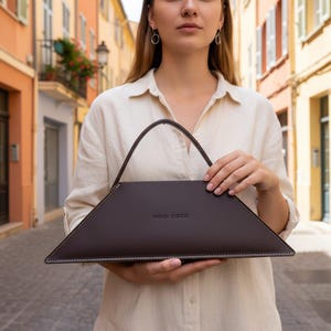 Puede incluir: Un bolso de mano marrón oscuro en forma de trapecio con un asa triangular. El bolso tiene costuras blancas alrededor de los bordes y el texto "NOCI COCO" está impreso en la parte delantera. El fondo está borroso.