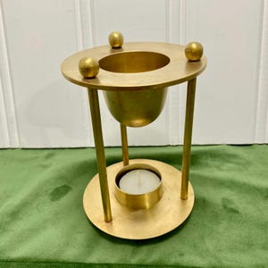 May include: A gold-coloured metal oil warmer featuring a circular top plate with three small spherical accents, a central bowl for oil, and three vertical support rods connecting to a circular base that holds a small white tea light candle.