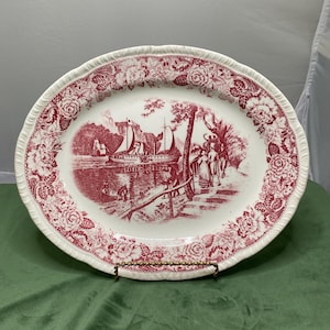 Peut inclure: Un plat de service ovale antique avec une bordure florale rouge et blanche et une scène de paysage centrale. Le paysage représente une rivière avec des voiliers, des personnages et des arbres. Le plat est soutenu par un support en métal doré.