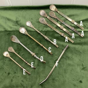 May include: A collection of nine metal bombilla straws for drinking mate, labelled with letters A through F. Each straw features a perforated spoon-shaped filter at one end and a long, slender stem, with varying decorative details and finishes.