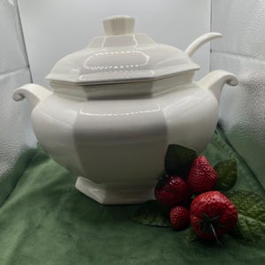 May include: A large, off-white ceramic soup tureen with a matching lid and integrated side handles. The tureen features an octagonal base and body design. A decorative cluster of three red strawberries with green leaves is positioned in the foreground.