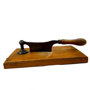 May include: An antique cheese slicer with a wooden base and a dark metal blade. The handle is made of wood and the slicer has a black metal support. The wooden base is rectangular and has a natural wood grain.