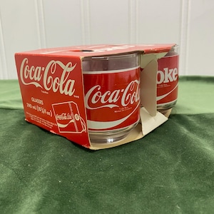 May include: A set of clear glass tumblers housed in a red cardboard carrier featuring the white Coca-Cola logo. The packaging displays the text Coca-Cola Glasses 290 ml (10 1/4 fl oz) and includes an official licensed product tag graphic.
