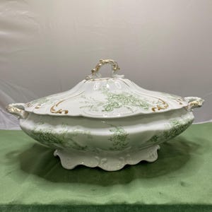 May include: White porcelain serving dish with a lid, featuring a green floral pattern and gold accents. The dish has a scalloped edge and ornate handles. A decorative piece for serving food.