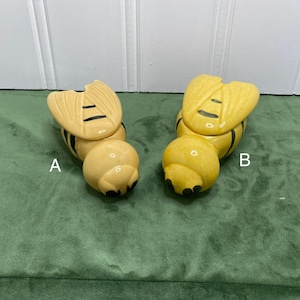 May include: Two ceramic bee-shaped trinket boxes. The bees are yellow with black stripes and black accents. The wings are a lighter yellow with black lines. The boxes are on a green surface.