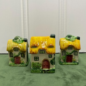 May include: Three ceramic house-shaped figurines featuring yellow thatched roofs, white walls, red doors, and green painted foliage. Each miniature building has small windows and a textured roof design, arranged in a row on a flat surface.