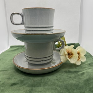 May include: Two stacked grey ceramic teacups with matching saucers. The cups have a subtle brown rim and a light grey glaze. The saucers are a slightly darker grey with a brown rim.
