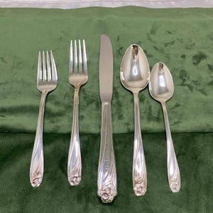 Peut inclure: Un ensemble de cinq couverts argentés, comprenant deux fourchettes, un couteau et deux cuillères, disposés sur une surface en velours vert. Les manches présentent un motif floral à la base. Le couteau porte une inscription sur le manche.
