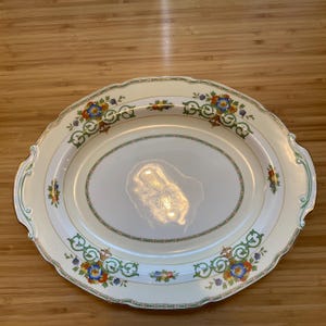 May include: An oval-shaped serving platter with a cream-colored background and a green, gold, and floral design. The platter has a raised rim and a slightly scalloped edge.