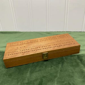 Könnte beinhalten: Ein rechteckiges Cribbage-Spielbrett aus Holz mit einer natürlichen, braunen Holzmaserung. Die Oberseite enthält mehrere Reihen gebohrter Löcher zum Zählen der Punkte. Ein kleiner, messingfarbener Metallverschluss ist vorne mittig angebracht.