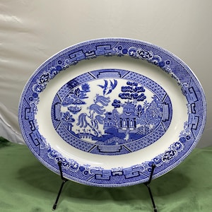May include: An oval ceramic platter featuring a blue and white willow pattern. The central design depicts a landscape with a pagoda, a willow tree, two birds in flight, and a small boat on water. The rim has a decorative geometric border.