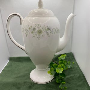 May include: White ceramic coffee pot with a floral design in shades of green. The pot has a lid, a handle, and a base. It is sitting on a green surface.