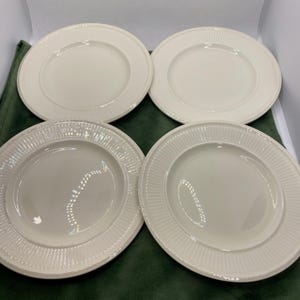 Könnte beinhalten: Set aus vier weißen Keramik-Dinnertellern mit erhöhtem Rand und einem dezenten Rippenmuster. Die Teller sind quadratisch auf einer grünen Oberfläche angeordnet.