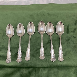 May include: Six silver spoons displayed on a green surface. Each spoon features an ornate handle design and a polished, reflective surface. The spoons are likely for serving or eating, with a classic aesthetic.