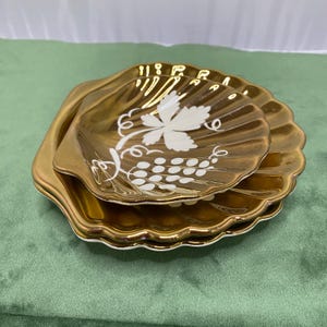 May include: A set of three gold-coloured serving dishes. The top dish is shell-shaped and features a white grape and leaf design. The other two dishes are square with scalloped edges. The dishes are on a green surface.