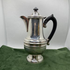 May include: A silver coffee pot with a black handle and a dark grey finial on the lid. The pot has a spout and a pedestal base. The pot is on a green surface.