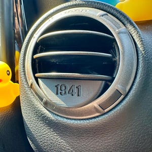 Könnte beinhalten: Nahaufnahme eines schwarzen Lederausstattungs eines Autos mit einem silbernen Lüftungsgitter. Das Lüftungsgitter hat ein kreisförmiges Design mit einem schwarzen Ring am Rand und einer schwarzen Mitte mit der Nummer "1941" darauf gedruckt.