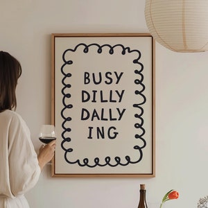 May include: A white framed print with black text that reads "Busy Dilly Dally Ing" in a decorative font. The print is hanging on a white wall above a table with a bottle of wine, a glass of wine, and a bowl of fruit.