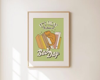 May include: A framed print with a light green background features a cartoon illustration of a sleeping orange cat resting on a stack of books. The text "It's Okay To Have A Bad Day" is written in a retro font.
