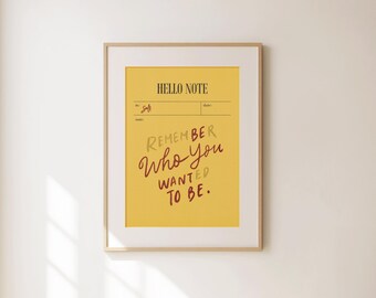Może przedstawiać: Oprawiony wydruk z żółtym tłem i napisem "HELLO NOTE" u góry. Tekst "REMEMBER Who you WANTED TO BE" jest napisany mieszanką brązu i złota. Rama ma jasny kolor drewna.