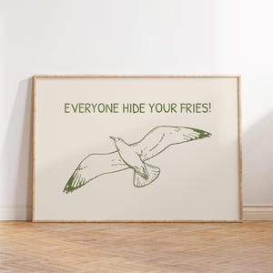 Pode incluir: Uma impress&atilde;o emoldurada com uma ilustra&ccedil;&atilde;o verde de uma gaivota em voo. O texto "EVERYONE HIDE YOUR FRIES!" est&aacute; impresso acima. O fundo &eacute; bege claro e a moldura &eacute; de madeira clara.