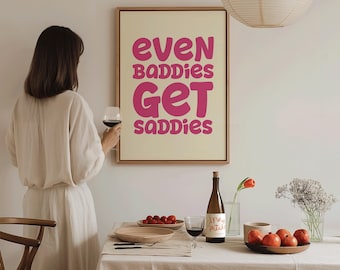 May include: A framed print with the text "EVEN BADDIES GET SADDIES" in pink letters. A woman in a cream-colored dress holds a glass of red wine. A table is set with a bottle of wine, fruit, and flowers.