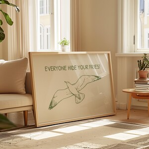 Pode incluir: Uma impress&atilde;o art&iacute;stica emoldurada com uma ilustra&ccedil;&atilde;o verde de uma gaivota em voo. O texto "EVERYONE HIDE YOUR FRIES!" est&aacute; impresso acima do p&aacute;ssaro. A obra de arte est&aacute; em uma moldura de madeira clara, encostada em uma parede.