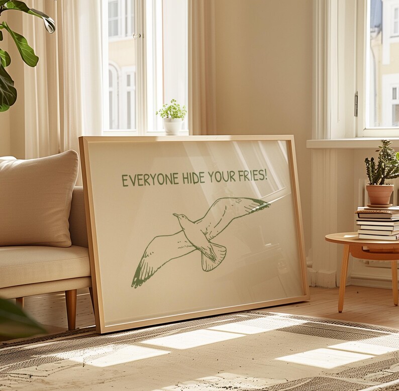 Pode incluir: Uma impress&atilde;o art&iacute;stica emoldurada com uma ilustra&ccedil;&atilde;o verde de uma gaivota em voo. O texto "EVERYONE HIDE YOUR FRIES!" est&aacute; impresso acima do p&aacute;ssaro. A obra de arte est&aacute; em uma moldura de madeira clara, encostada em uma parede.