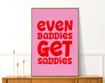 May include: A framed pink art print with the words "EVEN BADDIES GET SADDIES" in bold red letters. The artwork is displayed on a wooden cabinet with gold candle holders.