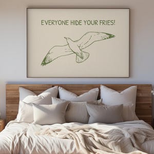 Pode incluir: Uma impress&atilde;o emoldurada com uma ilustra&ccedil;&atilde;o verde de uma gaivota em voo e o texto "EVERYONE HIDE YOUR FRIES!". A obra de arte est&aacute; acima de uma cama com roupa de cama em tons neutros.