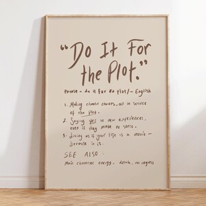 May include: A framed print featuring the phrase "Do It For the Plot" in a brown handwritten style. The print includes a definition and three points about the phrase. The frame is light wood, and the background is a beige colour.