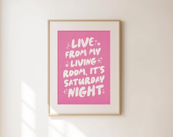 以下が含まれることがあります： ピンク色の背景に「LIVE FROM MY LIVING ROOM, IT'S SATURDAY NIGHT」と書かれた白い文字がプリントされた額入りのアートプリント。 プリントは薄茶色のフレームに入っており、白い壁に飾られています。