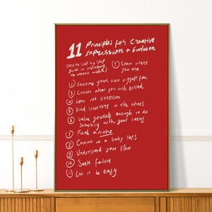 May include: A framed red art print with white handwritten text. The text lists "11 Principles for Creative Expression + Evolution" and includes phrases like "Find structure in the chaos" and "Fuck a niche". The frame is gold.