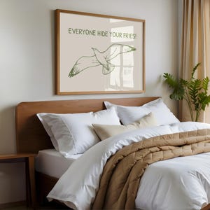 Pode incluir: Uma impress&atilde;o emoldurada com o texto "EVERYONE HIDE YOUR FRIES!" e uma ilustra&ccedil;&atilde;o verde de uma gaivota em voo. A impress&atilde;o est&aacute; acima de uma cama com roupa de cama branca e uma colcha acolchoada castanha. Uma pequena mesa de cabeceira de madeira tamb&eacute;m &eacute; vis&iacute;vel.